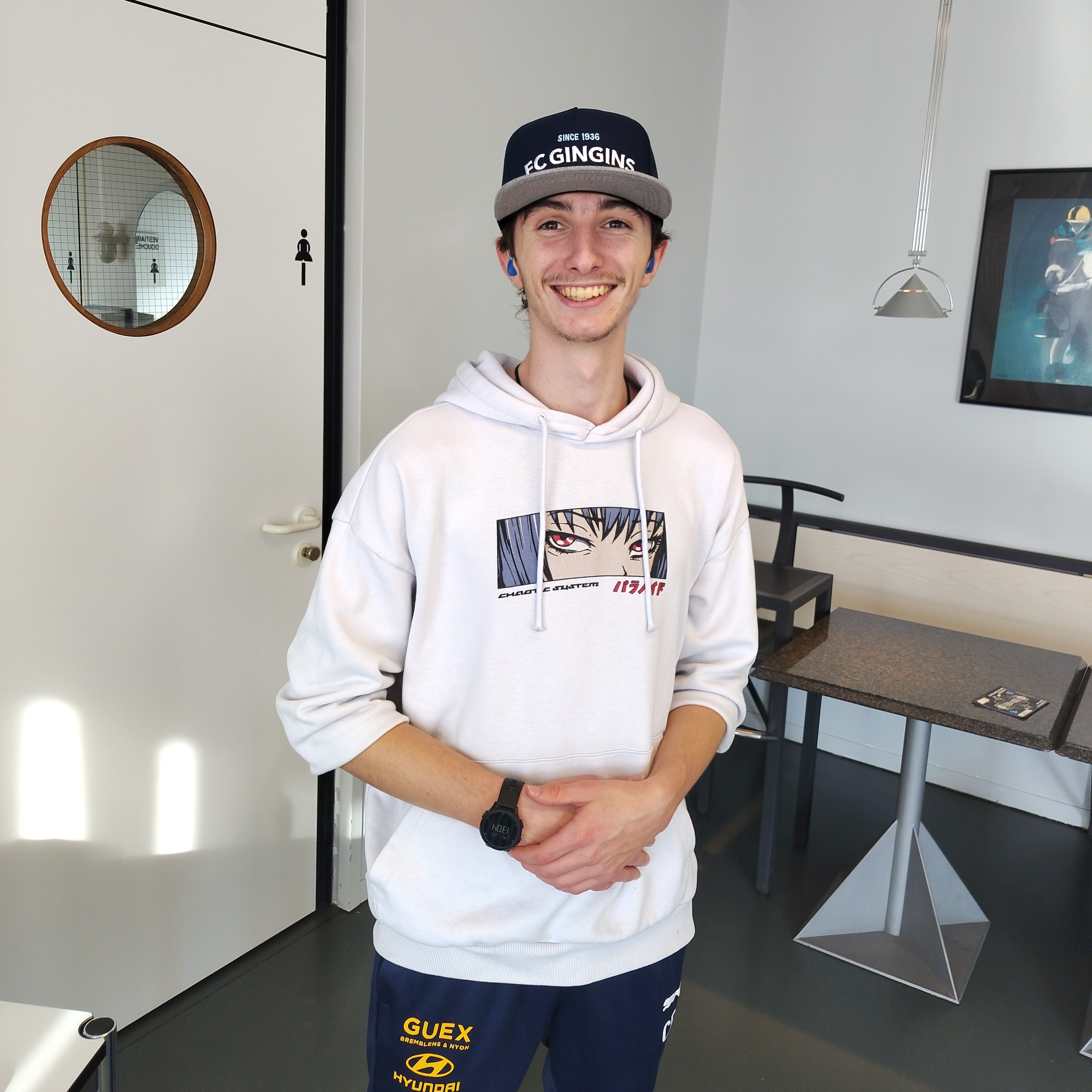 c'est moi, un jeune homme de 17 ans avec une petite moustache et un début de barbe, je porte un pull blanc avec des yeux d'un personnage de manga, j'ai également un training synthétique de foot bleu et je porte une casquette, de mon club de foot, qui est bleue avec écrit dessus : FC GINGINS 1936