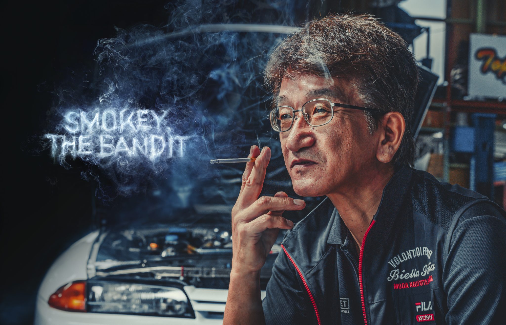 Kazuhiko nagata qui tien une cigarette dans sa main et la fumée écrit 'SMOKEY THE BANDIT' avec, en arrière plan, une toyota supra blanche qui a le capot ouvert pour qu'on voit le moteur 
